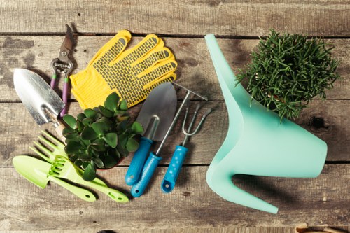 A collection of gardening tools and accessories arranged on a weathered wooden surface, including a pair of yellow gardening gloves, a small hand trowel, a cultivator, and pruning scissors, alongside a potted evergreen shrub and a light turquoise watering can. The tools are made of metal and plastic with blue handles, positioned in a casual manner. The wooden surface suggests an outdoor workspace, with natural light illuminating the scene, highlighting the textures of the tools and the greenery. This setup reflects typical gardening equipment used for outdoor landscaping and maintenance, suitable for services such as garden planting, pruning, and lawn care offered by companies like Landscapers Battersea. The overall composition emphasizes practical gardening tools in a natural outdoor environment, suitable for local clients in Battersea or nearby areas seeking professional gardening or landscaping solutions.