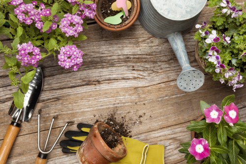 The image shows a wooden garden table with gardening tools and decorative pots. There is a small terracotta pot filled with soil and a pair of yellow gardening gloves placed nearby. A garden fork with a wooden handle and metal tines rests on the table, along with a hand rake and a small gardening trowel. Several pots contain vibrant flowering plants with pink, purple, and white flowers, adding colour to the scene. A grey watering can with a textured surface is positioned at the top right, and the table surface displays natural wood grain. This outdoor space appears well-maintained, suitable for gardening and landscaping activities, consistent with services offered by Battersea landscapers specializing in garden design and maintenance.