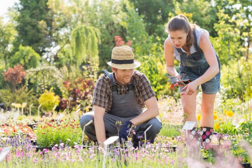 A gardener and a young girl are working together in a vibrant garden during daylight hours, with lush green trees and colourful flowering plants in the background. The gardener, wearing a checkered shirt, grey apron, and a straw hat, is kneeling on the grass and tending to small plants or flowers in a flowerbed. The girl, dressed in a sleeveless top and denim shorts, is standing beside the gardener, holding garden shears and assisting with the planting or trimming process. The garden features a well-maintained lawn with dense, healthy grass, bordered by a variety of flower beds containing pink, purple, and yellow blooms. In the distance, a mix of tall trees and shrubs provide natural shade and background greenery, creating a peaceful outdoor environment suitable for gardening activities. The scene captures a bright, sunny day with natural light illuminating the garden, and the overall setting aligns with professional gardening and landscaping services offered by Landscapers Battersea in the Battersea area, integrating natural plant growth, garden layout, and outdoor maintenance features.