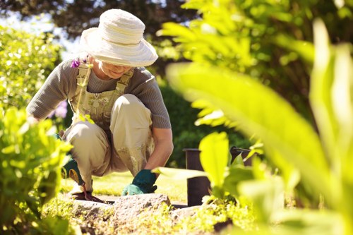 A woman wearing a large sun hat, glasses, and gardening gloves is kneeling on a lush green lawn in a landscaped garden in Battersea. She appears to be planting or tending to a flower bed bordered by small brown bricks. The garden features a well-maintained grass area with dense, vibrant foliage and bushes surrounding her, providing shade and a natural backdrop. In the background, there are taller trees and more greenery, creating a peaceful outdoor environment. The weather is sunny with bright natural light illuminating the scene, and the overall setting suggests careful garden maintenance and landscaping work typical of professional gardening services in the Battersea area. The scene emphasizes outdoor lawn care, planting, and garden upkeep, aligning with gardening service activities provided by Landscapers Battersea.