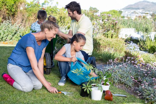 A family of four working together in a lush backyard garden during daylight, with a neatly maintained grass lawn in the foreground and an array of flowering plants and shrubs along the garden borders. The mother is kneeling on the grass, planting or tending to a small plant, while the young girl next to her is sitting on the ground, holding a small gardening fork and a blue garden bag filled with gardening tools and supplies. The father and a young boy are standing behind, observing or assisting with gardening tasks in the background. The garden features a mix of vibrant flowering plants, variegated leaves, and green shrubbery, with a wooden garden edge or border visible. Natural sunlight illuminates the scene, suggesting a bright, clear day. The scene reflects typical outdoor garden maintenance and family gardening activities, aligned with landscaping services in Battersea, with a focus on healthy, well-kept outdoor spaces.