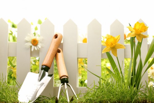 A close-up view of a small, well-maintained garden area in Battersea featuring a white picket fence in the background, with vibrant yellow daffodils and white daisies growing along the base of the fence. The foreground includes a patch of lush, green grass with a slightly uneven texture, indicative of regular lawn care. Resting against the fence are garden tools, including a hand trowel and pruning shears with wooden handles, suggesting ongoing gardening activities. The scene is illuminated by natural daylight under clear weather conditions, creating bright highlights and natural shadows on the plants and tools. The overall setting depicts a tidy, well-kept outdoor space suitable for landscaping, garden maintenance, and outdoor beautification services offered by Landscapers Battersea, aligning with the theme of garden renovation and landscaping in the Battersea area.