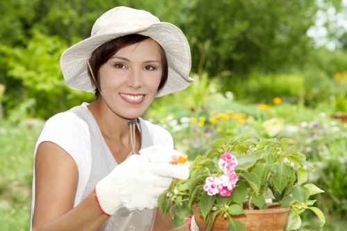 The image shows a woman in a white wide-brimmed hat and gardening gloves tending to a potted flowering plant in a well-maintained garden. She is smiling and appears to be engaged in gardening activities, with colorful flowers and lush green foliage in the background. The garden features a grassy lawn, flower beds with various plants, and trees that provide shade. The scene suggests a healthy, organized outdoor space typical of residential landscaped gardens in Battersea, with natural daylight indicating a bright, clear day. The woman’s focus on the plant reflects attentive gardening care, supported by professional gardening services from Landscapers Battersea that help maintain vibrant and inviting outdoor environments, including flower bed planting and lawn care.