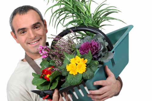 The image depicts a smiling man with short dark hair holding a green plastic garden tool caddy filled with a variety of vibrant flowering plants, including purple, yellow, and red blossoms, and lush green foliage. Behind him, a potted shrub with tall, slender green leaves is visible, and the background appears to be an outdoor garden setting with natural daylight. The scene is clean and well-maintained, emphasizing gardening and outdoor care activities typical of landscaping services in Battersea. The overall environment suggests a well-tended garden area, suitable for professional gardening and landscaping work by companies like Landscapers Battersea. The image showcases elements relevant to gardening tasks such as planting, plant care, and garden maintenance, aligning naturally with services offered in the Battersea area, SW11 postcode, in a professional and approachable manner.