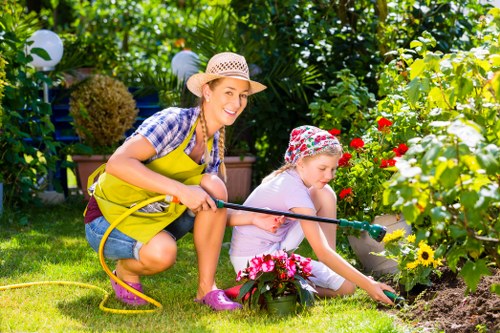 A woman and a young girl gardening in a well-maintained front garden, with lush green foliage and a variety of colorful flowering plants, including red, yellow, and pink blooms. The woman, wearing a straw sun hat, a blue checkered shirt, and a yellow apron, is kneeling with a smile while holding a garden hose, watering the plants. The girl, dressed in a floral sun hat and pink shoes, is focused on tending to a flower bed, carefully placing or adjusting flowers among the soil and existing plants. The garden features a neatly mown lawn, decorative hanging lanterns, and well-defined flower borders, creating a bright and inviting outdoor space typical of residential gardens in Battersea. The scene is illuminated by natural sunlight, suggesting a warm and sunny day, and reflects a caring outdoor environment suitable for professional gardening services aimed at enhancing garden health and visual appeal.
