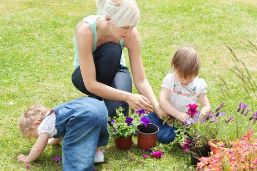 A woman with short blonde hair, dressed in a light blue top and dark jeans, is kneeling on a well-maintained grassy lawn in a garden, engaging in planting or tending to potted plants. Two young children, one with blonde hair wearing a white shirt and denim overalls, and the other with light brown hair in a floral dress, are also on the lawn; the child in overalls is bending forward, possibly inspecting or picking flowers, while the other is sitting and handling vibrant pink and purple flowers in a black pot. The garden features lush, bright green grass, with flower beds containing colorful blooms such as pink, purple, and orange flowers in containers and directly in the ground. In the background, there are trees and shrubs, creating a natural outdoor environment suitable for family gardening activities. The scene appears to be during daytime with soft natural lighting, typical of mild weather, and promotes outdoor family gardening activities, aligning with services offered by Landscapers Battersea in the Battersea area.