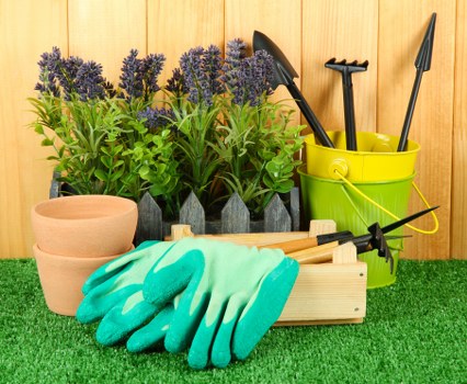 The image features a small outdoor garden space with a well-maintained lawn area in the foreground, covered with vibrant green artificial grass. Behind the lawn, there is a collection of potted plants with purple flowering lavender and bright green foliage, arranged along a wooden fence or wall. To the right, a yellow plastic bucket holds various gardening tools such as a trowel, transplanter, and hand rake, all with black handles. In front of these, a pair of gardening gloves in a bright blue-green color rest on a natural wooden tool tray, which also contains a small garden fork and trowel. The overall setting appears to be a tidy, organized outdoor garden corner designed for gardening and maintenance activities, with natural light illuminating the scene, suggesting a clear, mild weather day. This setup reflects typical tools and plant arrangements used in landscaping and garden upkeep, relevant to services offered by Landscapers Battersea and supporting sustainable gardening practices in the local area of Battersea, London postcode SW11.