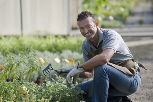 A smiling man wearing a grey shirt, jeans, and gardening gloves is kneeling on a gravel path in a lush garden, holding a small garden rake. Behind him, there are neatly maintained flower beds with various green foliage and some flowering plants. The garden features a well-kept lawn in the foreground, bordered by planting beds and a wooden fence in the background, suggesting an outdoor space typical of residential gardens in Battersea. The scene is illuminated by natural daylight, highlighting the vibrant greenery and natural tones of the garden environment, with a slightly blurred background emphasizing the gardener's focus on the plants. This setting underscores professional gardening services, such as planting, weeding, and general outdoor maintenance, that a company like Landscapers Battersea might provide for local clients looking to enhance their garden’s appearance and health.